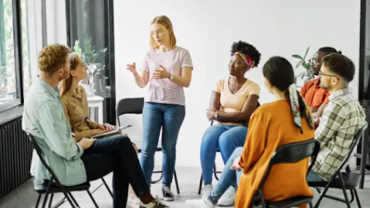 Grupo de personas sentadas en círculo mientras una mujer interviene de pie.